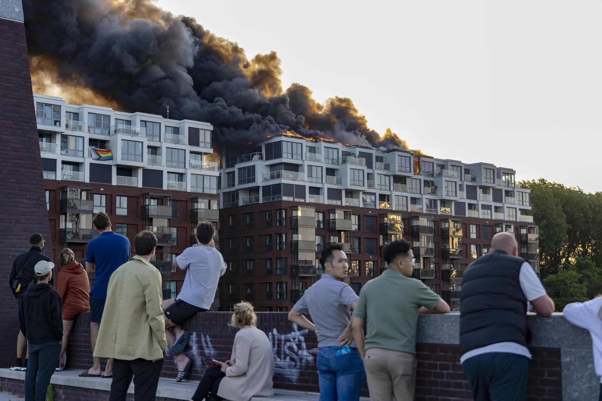 Brand met zware rookontwikkeling op het dak van een appartementencomplex, bekeken door omstanders.