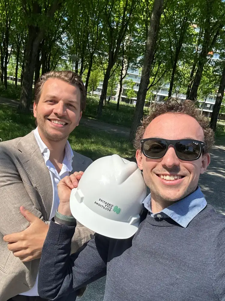 Twee collega's buiten tussen de bomen, een van hen met een bouwhelm in de hand