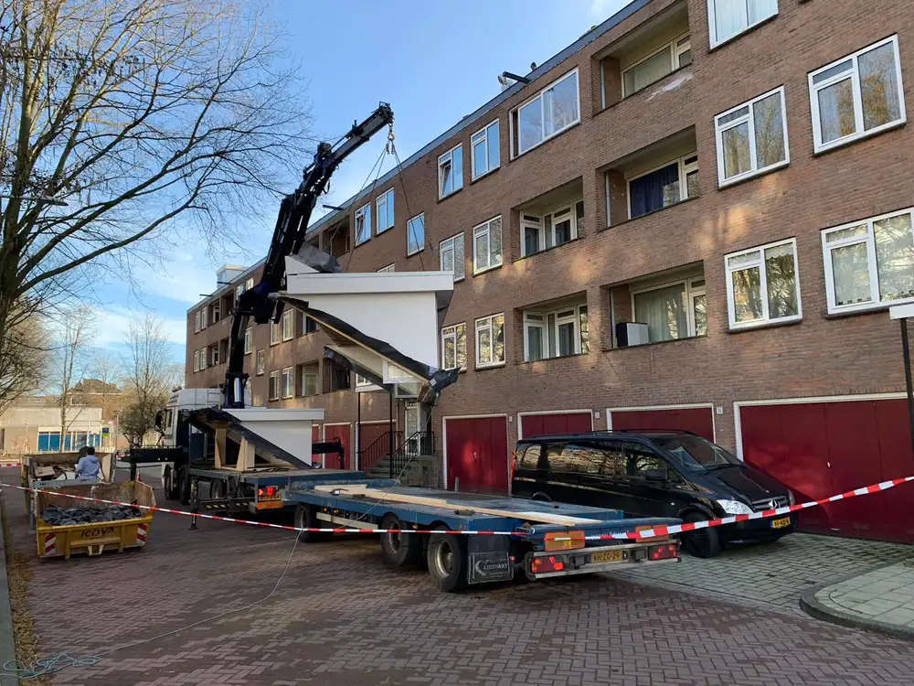Een kraan hijst een prefabelement op het dak van een woongebouw, gezien vanaf de straat
