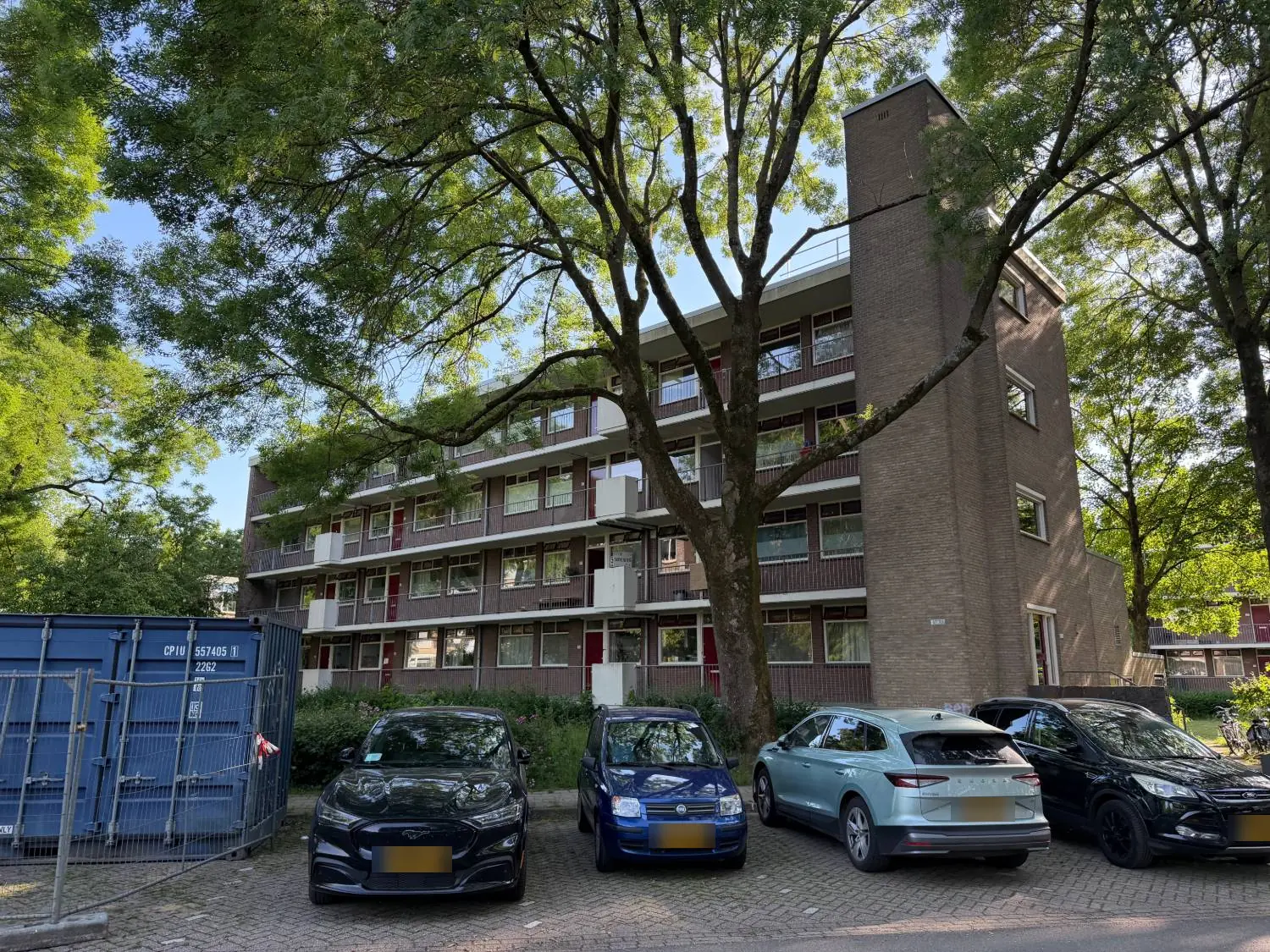 Galerijflat van vier woonlagen met een bakstenen trappenhuis, achter een grote boom en geparkeerde auto's