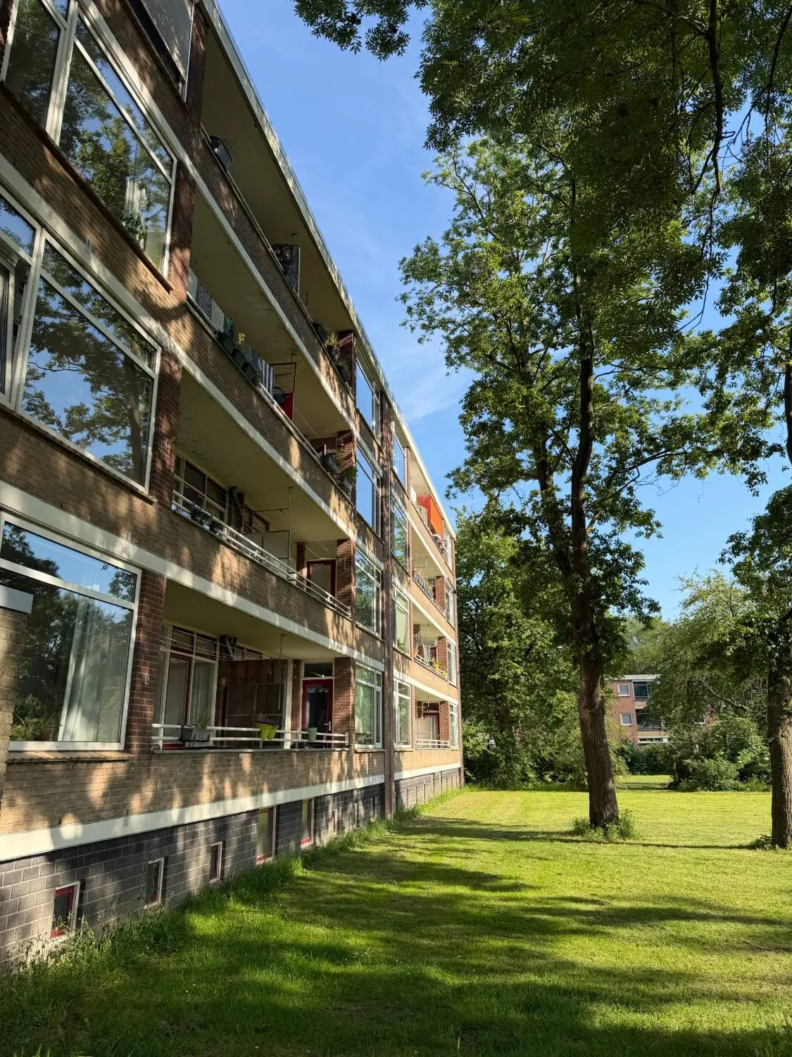 Bakstenen flat met balkons op elke verdieping, langs een grasveld met hoge bomen