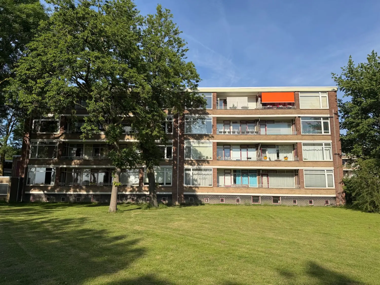 Bakstenen flat van vier woonlagen met balkons en een oranje zonnescherm, achter een grasveld met twee bomen