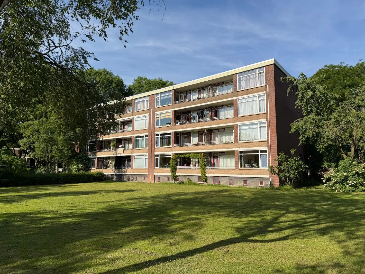 Bakstenen flat van vier woonlagen met balkons en grote ramen, aan een grasveld tussen bomen